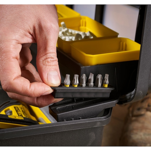 Close-up of a hand picking up a set of screwdriver bits from a black and yellow organizer box, with several compartments and tools visible.