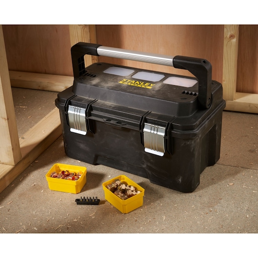 A black STANLEY toolbox with a metal handle and two metal latches, placed on a workshop floor. In front of the toolbox are two small yellow organizer trays containing various fittings and bits.
