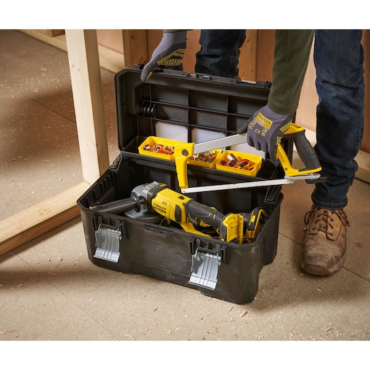 A black STANLEY toolbox with metal latches is open on the floor. Inside are various tools and hardware, including a yellow and black power drill, a hacksaw, and small compartments holding screws and fittings. A person wearing gloves and work boots is organizing the tools.