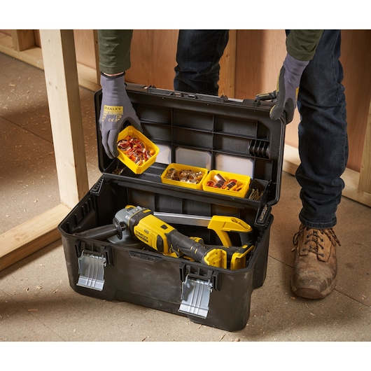 A person wearing gloves is opening a STANLEY tool box filled with various small parts and a yellow power tool, positioned in a workspace with wooden framing.