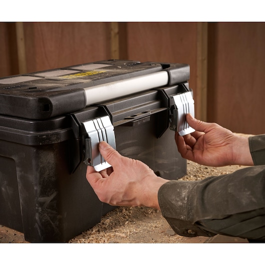 A person is opening the sturdy metal latches of a black STANLEY toolbox on a dusty workbench in a workshop.