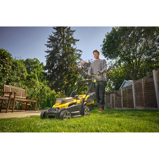 Photo of STANLEY SCMWP3401M1-XE lawn mower in use on green grass with wooden fence and bench in background.