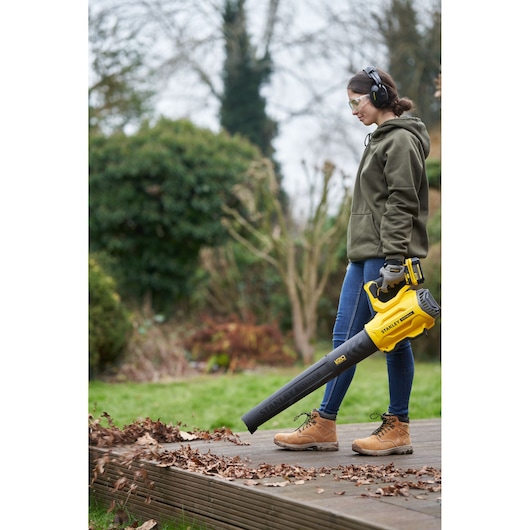 Photo of a person using a STANLEY SFMCBL7M1 cordless leaf blower, yellow and black, clearing leaves in a garden.