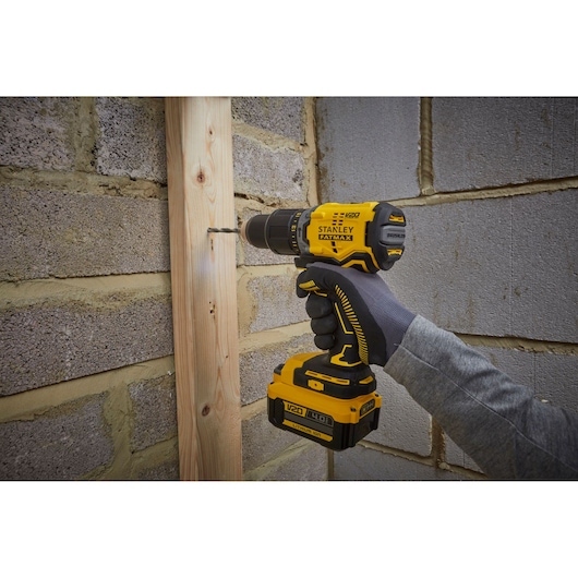 Stanley SFMCD715M12 cordless drill being used to drill wood against a brick wall, close-up view.