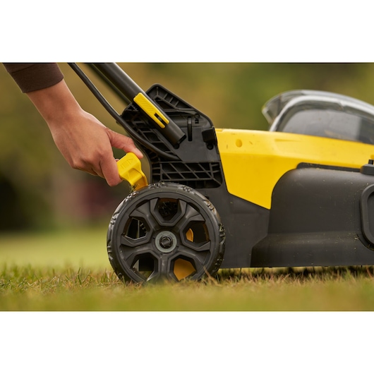 STANLEY SFMCMW2651M lawn mower side view, hand adjusting yellow wheel height lever outdoors.