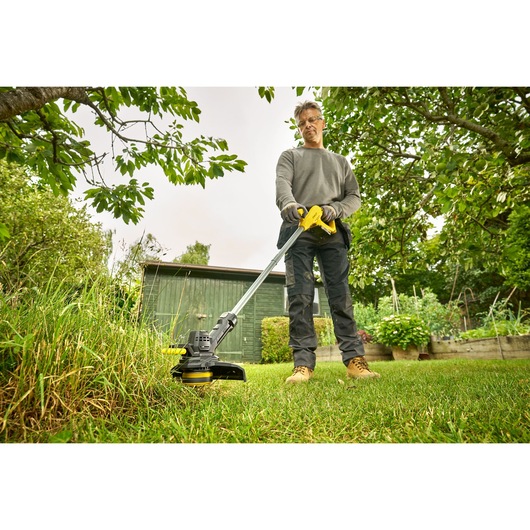 STANLEY SFMCST925M1 grass trimmer in use, person trimming lawn in garden with green shed in background.