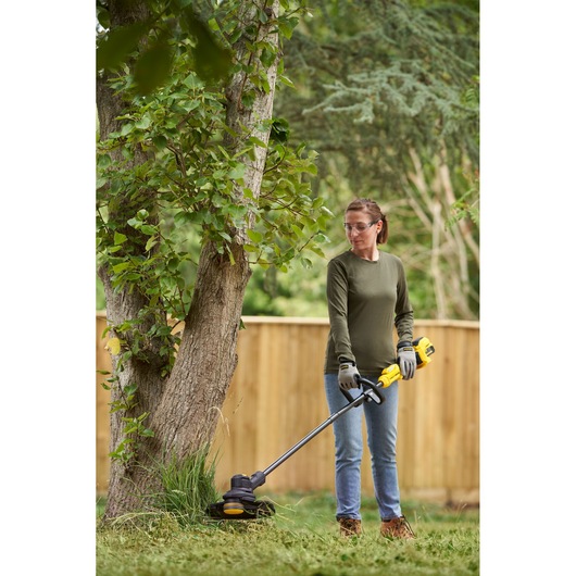 Photo of person using STANLEY SFMSTB930M grass trimmer outdoors near a tree, wearing gloves and casual clothing.