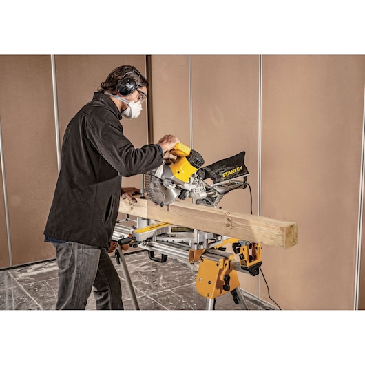 Photo of a person using a STANLEY SM18-B2C miter saw to cut a wooden beam indoors, wearing safety gear.