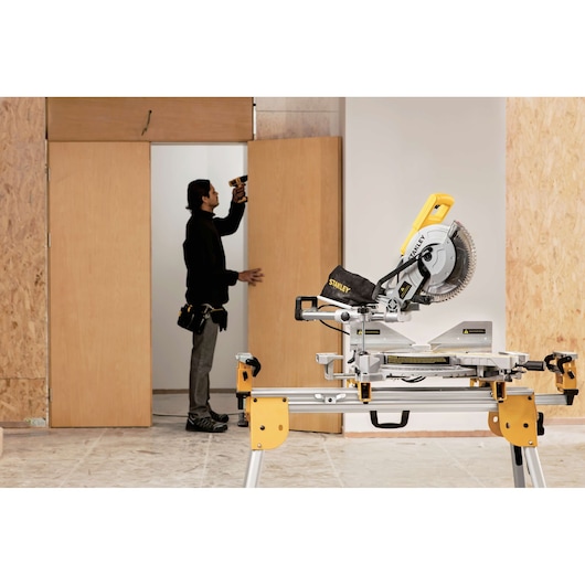 STANLEY SM18-B2C miter saw on stand in workshop, person using drill near wooden door in background.