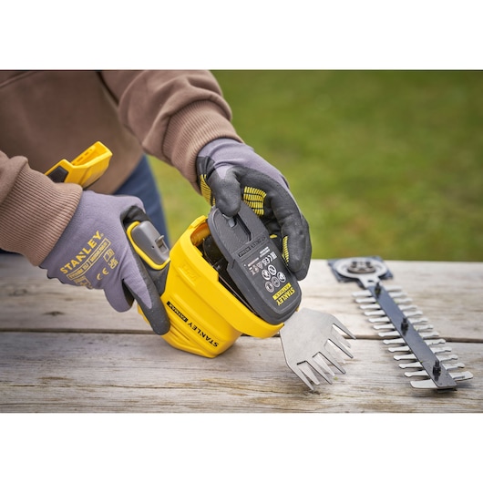 STANLEY STCSS20D1 cordless shrub and grass shear being fitted with battery, gloves worn, parts on wooden table.