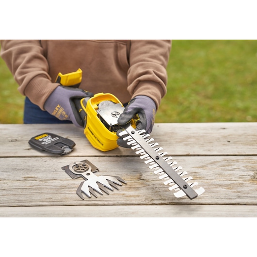 STANLEY STCSS20D1 hedge trimmer in use, with blade attachment being installed, battery and extra blade visible on table.