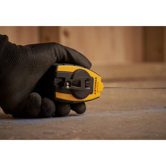 Photo of STANLEY STHT0-47147 chalk line reel held in a gloved hand, yellow and black, marking a surface.