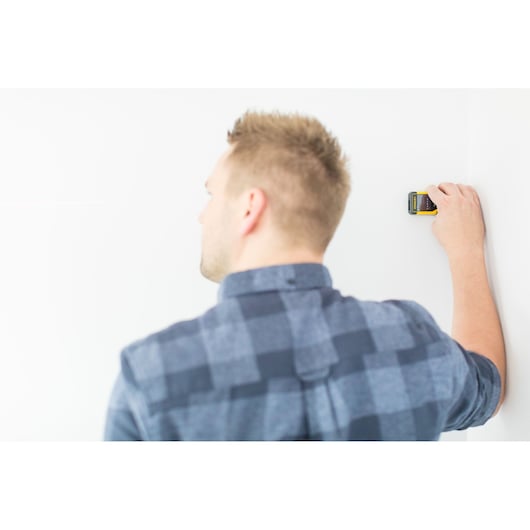 Photo of STANLEY STHT1-77032 stud finder held against a white wall by a person in a blue plaid shirt.