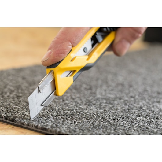STANLEY STHT10266-0 utility knife cutting grey carpet, close-up photo, yellow and black handle visible.