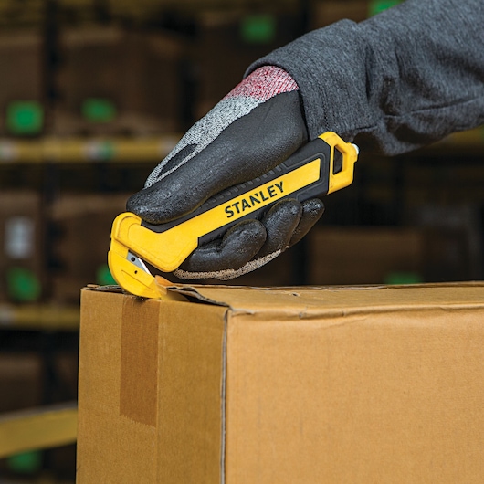 Photo of STANLEY STHT10356 safety utility knife cutting brown cardboard box in warehouse setting.