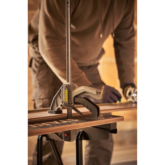 STANLEY STST83400-1 bar clamp securing wood on workbench, person working in background, side view.