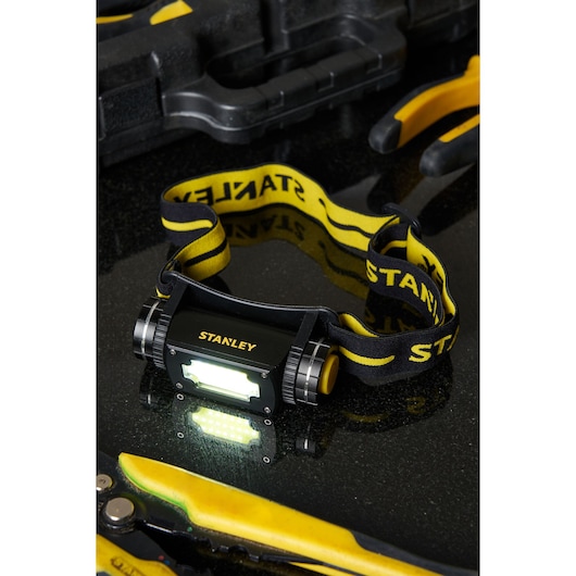 STANLEY SXLS50100E headlamp with yellow-black strap, shown illuminated on black surface with tools nearby.