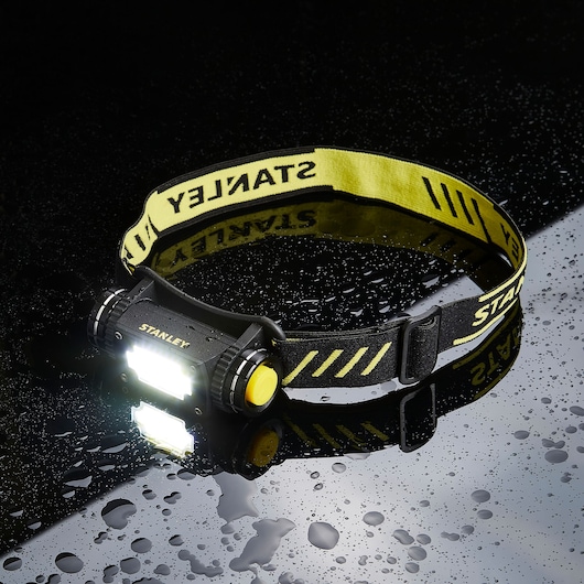 STANLEY SXLS50100E headlamp with yellow-black strap, LED light on, shown on wet surface.
