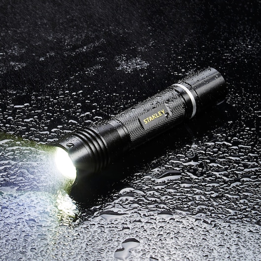 Stanley SXLS50104E waterproof black flashlight on wet surface, emitting bright light, side angle.