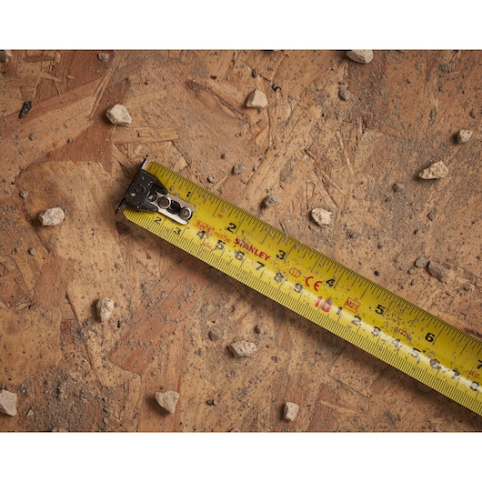 STANLEY XTHT0-33503 measuring tape close-up on a dusty wooden surface with small rocks.